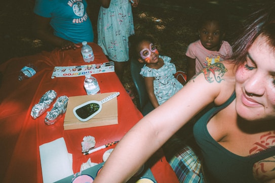 Several people, including children, are gathered around a table covered with a red cloth. The table has bottles of water, wrapped food items, a dish with a ladle, and some papers. One child has face paint featuring colorful designs. An adult with tattoos is smiling at the right front of the scene.