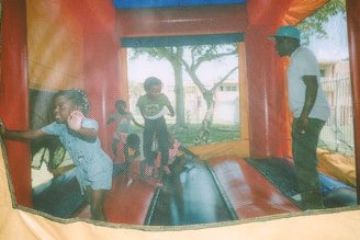 Several children and an adult are playing inside a colorful inflatable bounce house. The joyful atmosphere is evident as the children move energetically. The bounce house is situated outdoors, with trees visible in the background.