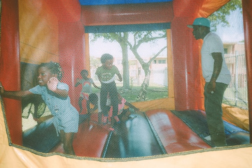 Several children and an adult are playing inside a colorful inflatable bounce house. The joyful atmosphere is evident as the children move energetically. The bounce house is situated outdoors, with trees visible in the background.
