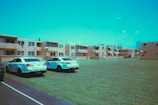 Two police cars are parked on a grassy area in front of a group of beige apartment buildings. The sky appears bright, and there are a few clouds visible. The buildings have multiple satellite dishes attached, and the scene conveys an urban neighborhood setting.