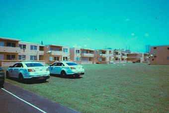 Two police cars are parked on a grassy area in front of a group of beige apartment buildings. The sky appears bright, and there are a few clouds visible. The buildings have multiple satellite dishes attached, and the scene conveys an urban neighborhood setting.