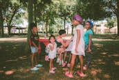 A group of children and one adult are in a park surrounded by trees. The children are standing and smiling, while the adult is seated at a portable table. The scene is lit by natural daylight, with dappled sunlight filtering through the branches.