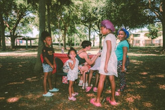 A group of children and one adult are in a park surrounded by trees. The children are standing and smiling, while the adult is seated at a portable table. The scene is lit by natural daylight, with dappled sunlight filtering through the branches.