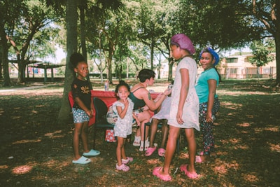 A group of children and one adult are in a park surrounded by trees. The children are standing and smiling, while the adult is seated at a portable table. The scene is lit by natural daylight, with dappled sunlight filtering through the branches.