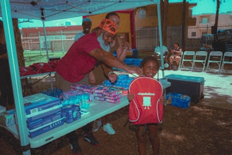 A smiling child holds a red Nickelodeon backpack while a man with a white beard and an orange cap gestures toward them. The setting is an outdoor tent with tables displaying various packaged goods and supplies. Three other individuals are in the background, with folding chairs lined up in the right corner. Natural light and shadows create a relaxed atmosphere.