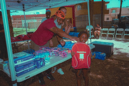 A smiling child holds a red Nickelodeon backpack while a man with a white beard and an orange cap gestures toward them. The setting is an outdoor tent with tables displaying various packaged goods and supplies. Three other individuals are in the background, with folding chairs lined up in the right corner. Natural light and shadows create a relaxed atmosphere.