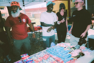 Several people are standing under a canopy at an outdoor event, appearing to engage in conversation. A table in front of them is filled with various packaged goods, including hygiene products like toothpaste and toothbrushes. The background shows a fenced area with grass and a building.