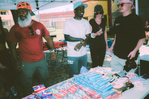 Several people are standing under a canopy at an outdoor event, appearing to engage in conversation. A table in front of them is filled with various packaged goods, including hygiene products like toothpaste and toothbrushes. The background shows a fenced area with grass and a building.