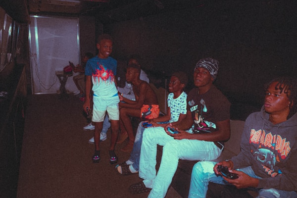 A group of people are sitting together in a dimly lit room, engrossed in playing video games. Several individuals are holding game controllers, and the space has a relaxed atmosphere. The room is dark, with light coming from screens and a light source from a nearby open door or window.