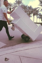 A person in a white shirt is maneuvering a large white furniture piece on a wheeled dolly across a street. Palm trees are visible in the background, suggesting a warm climate. The scene has a vintage filter effect.