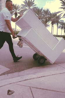 A person in a white shirt is maneuvering a large white furniture piece on a wheeled dolly across a street. Palm trees are visible in the background, suggesting a warm climate. The scene has a vintage filter effect.