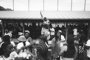 A group of people gather in an outdoor pavilion, engaged in lively social interaction. One person stands on a table with an arm raised triumphantly while others around him are either talking, smiling, or looking towards the activity. Some individuals wear casual summer clothing, including hats and basketball jerseys. The pavilion roof is metal, and there are more people in the shaded area beneath it.