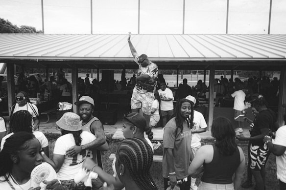 A group of people gather in an outdoor pavilion, engaged in lively social interaction. One person stands on a table with an arm raised triumphantly while others around him are either talking, smiling, or looking towards the activity. Some individuals wear casual summer clothing, including hats and basketball jerseys. The pavilion roof is metal, and there are more people in the shaded area beneath it.