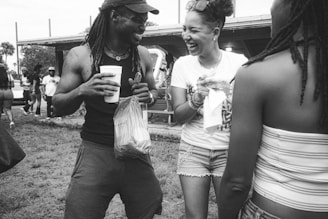 A group of people are outdoors, engaging in lively conversation and laughter. One person holds a drink and a plastic bag, while another is smiling broadly, creating an atmosphere of joy and camaraderie.