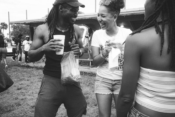 A group of people are outdoors, engaging in lively conversation and laughter. One person holds a drink and a plastic bag, while another is smiling broadly, creating an atmosphere of joy and camaraderie.