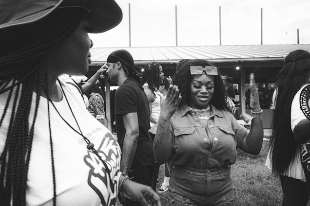 A group of people are gathered outdoors during the day. One woman in focus is gesturing with both hands in front of her, wearing a jumpsuit and sunglasses on her head. Other individuals in casual attire, including someone wearing a hat and a necklace, are interacting around her. The background includes a sheltered structure and an open field.