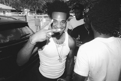 A black and white photograph featuring a smiling person in the foreground, holding up a peace sign with fingers. The individual wears a tank top and a necklace, with additional people in the background engaged in conversation. The setting appears to be outdoors, possibly in a residential area with visible fence and parked cars.