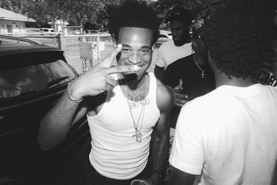 A black and white photograph featuring a smiling person in the foreground, holding up a peace sign with fingers. The individual wears a tank top and a necklace, with additional people in the background engaged in conversation. The setting appears to be outdoors, possibly in a residential area with visible fence and parked cars.
