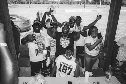 A group of people gathered under a wooden shelter, smiling and making celebratory gestures. They are wearing casual clothing, with some holding cups, and the setting appears to be outdoors with grass and trees in the background. There's a parked car visible on the left.