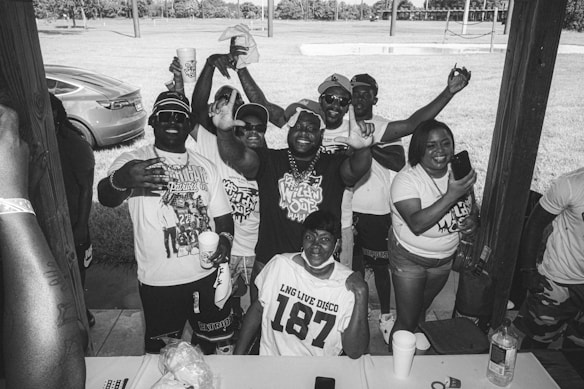 A group of people gathered under a wooden shelter, smiling and making celebratory gestures. They are wearing casual clothing, with some holding cups, and the setting appears to be outdoors with grass and trees in the background. There's a parked car visible on the left.
