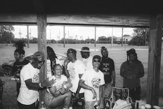 A group of people gather under a wooden shelter in an outdoor setting. They appear to be enjoying themselves, with some smiling and engaged in conversation. The background shows a grassy area with palm trees and sporting fields, while a car is partially visible. Most individuals are wearing casual clothing, some in white shirts with printed designs.