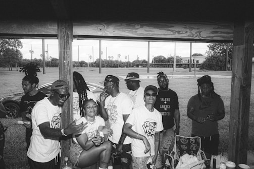A group of people gather under a wooden shelter in an outdoor setting. They appear to be enjoying themselves, with some smiling and engaged in conversation. The background shows a grassy area with palm trees and sporting fields, while a car is partially visible. Most individuals are wearing casual clothing, some in white shirts with printed designs.