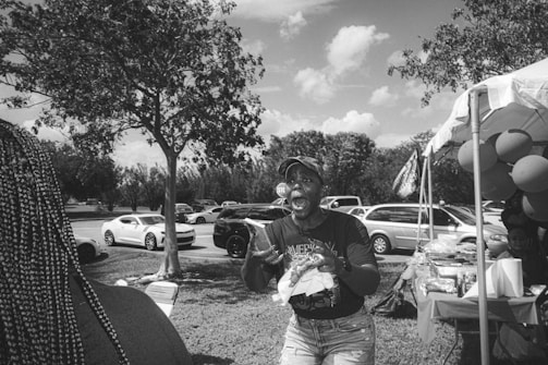 A lively outdoor scene featuring a person expressing excitement or joy. The image is black and white, showing cars parked in the background, a tree, and a canopy tent with various items on a table. Balloons are attached to the side of the tent.