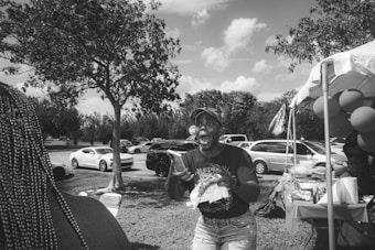 A lively outdoor scene featuring a person expressing excitement or joy. The image is black and white, showing cars parked in the background, a tree, and a canopy tent with various items on a table. Balloons are attached to the side of the tent.