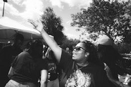 A group of people gathered outdoors, with one person in the foreground wearing sunglasses and reaching their hand out enthusiastically. The background includes trees and a tent, indicating a social or community event.