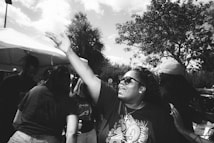 A group of people gathered outdoors, with one person in the foreground wearing sunglasses and reaching their hand out enthusiastically. The background includes trees and a tent, indicating a social or community event.