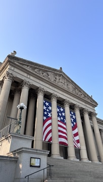 A neoclassical building with large columns and a detailed pediment, featuring a large American flag banner draped between the columns. The sky is clear and blue, enhancing the visibility of the structure's stone facade and decorative elements.