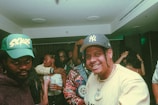 A group of people in a dimly lit room, likely at a party or gathering. Two men are in the foreground, both wearing caps with distinct logos. Others are seen mingling or dancing in the background. One person is smiling directly at the camera, suggesting a lively and friendly atmosphere.