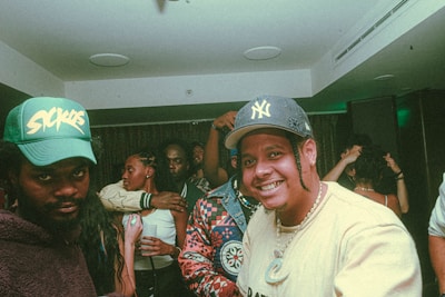 A group of people in a dimly lit room, likely at a party or gathering. Two men are in the foreground, both wearing caps with distinct logos. Others are seen mingling or dancing in the background. One person is smiling directly at the camera, suggesting a lively and friendly atmosphere.