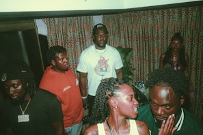 A group of people are gathered indoors, wearing casual clothing. Each person is engaged in either conversation or standing quietly. The setting features patterned curtains and some indoor plants, suggesting a cozy room atmosphere.