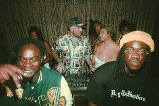 A group of people are gathered together in a dimly lit indoor setting. Three individuals are prominent in the foreground; one man on the left is smiling, while another person on the right appears more serious. The center individual is wearing a patterned shirt and is talking to a woman in a white top. There is DJ equipment in front of them, suggesting a party or nightclub atmosphere. The background features patterned curtains and more people engaging socially.