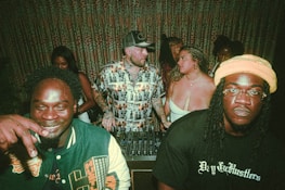 A group of people are gathered together in a dimly lit indoor setting. Three individuals are prominent in the foreground; one man on the left is smiling, while another person on the right appears more serious. The center individual is wearing a patterned shirt and is talking to a woman in a white top. There is DJ equipment in front of them, suggesting a party or nightclub atmosphere. The background features patterned curtains and more people engaging socially.