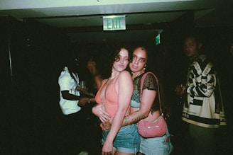 Two people pose closely together in a dimly lit setting, with one wearing a coral top and denim shorts and the other in a black mesh top with a pink bag. There are other individuals in the background, suggesting a social gathering or party atmosphere.