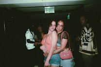 Two people pose closely together in a dimly lit setting, with one wearing a coral top and denim shorts and the other in a black mesh top with a pink bag. There are other individuals in the background, suggesting a social gathering or party atmosphere.