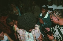A group of people are gathered together in a dimly lit setting, likely a social or party environment. They are dressed in casual attire, with some wearing distinctive patterns and colors. The atmosphere appears lively and energetic.