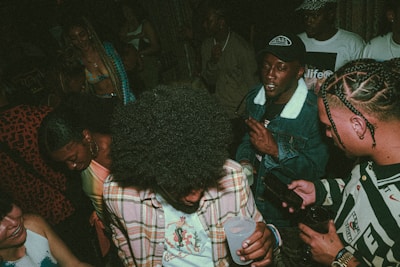A group of people are gathered together in a dimly lit setting, likely a social or party environment. They are dressed in casual attire, with some wearing distinctive patterns and colors. The atmosphere appears lively and energetic.