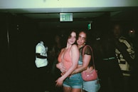 Two people are posing closely together in a dimly lit environment that appears to be a nightclub or party. They are wearing casual, fashionable clothing, with one person dressed in a tank top and shorts, and the other in a top with mesh sleeves and jeans. Other people are partially visible in the background, and there's an illuminated exit sign above.