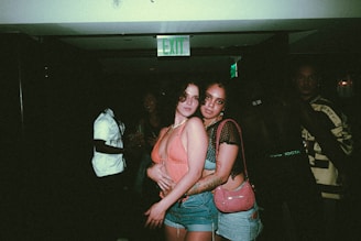 Two people are posing closely together in a dimly lit environment that appears to be a nightclub or party. They are wearing casual, fashionable clothing, with one person dressed in a tank top and shorts, and the other in a top with mesh sleeves and jeans. Other people are partially visible in the background, and there's an illuminated exit sign above.