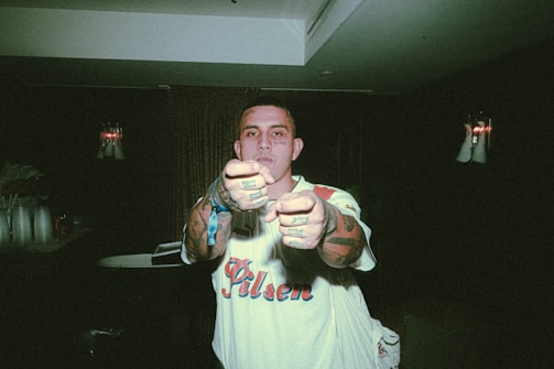 A person with tattoos and a shirt printed with 'Pilsen' stands indoors pointing fingers directly at the camera. The room is dimly lit with some ambient light visible in the background, which includes candles or wall sconces. The person's expression is serious and focused.