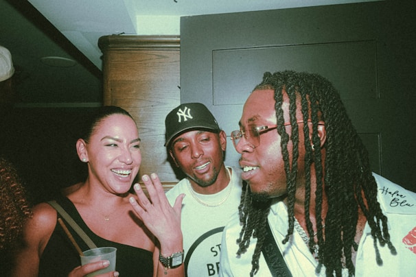 Three people are together indoors, enjoying a lively and friendly conversation. The woman is holding a drink and laughing, while the two men, wearing casual outfits, are engaged in the interaction. The atmosphere appears to be social and informal.