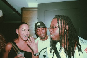 Three people are together indoors, enjoying a lively and friendly conversation. The woman is holding a drink and laughing, while the two men, wearing casual outfits, are engaged in the interaction. The atmosphere appears to be social and informal.