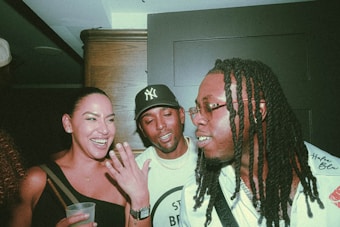 Three people are together indoors, enjoying a lively and friendly conversation. The woman is holding a drink and laughing, while the two men, wearing casual outfits, are engaged in the interaction. The atmosphere appears to be social and informal.