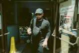 A person wearing dark clothing and a cap exits a building through glass doors. They are carrying a drink cup and wearing sunglasses. To the left, a chalkboard with colorful writing advertises 'El almacén Argentino.' There is a yellow caution sign on the floor, and to the right, a poster on the door promotes a restaurant named 'The Knife.'