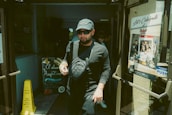 A person wearing dark clothing and a cap exits a building through glass doors. They are carrying a drink cup and wearing sunglasses. To the left, a chalkboard with colorful writing advertises 'El almacén Argentino.' There is a yellow caution sign on the floor, and to the right, a poster on the door promotes a restaurant named 'The Knife.'