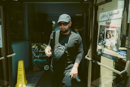 A person wearing dark clothing and a cap exits a building through glass doors. They are carrying a drink cup and wearing sunglasses. To the left, a chalkboard with colorful writing advertises 'El almacén Argentino.' There is a yellow caution sign on the floor, and to the right, a poster on the door promotes a restaurant named 'The Knife.'