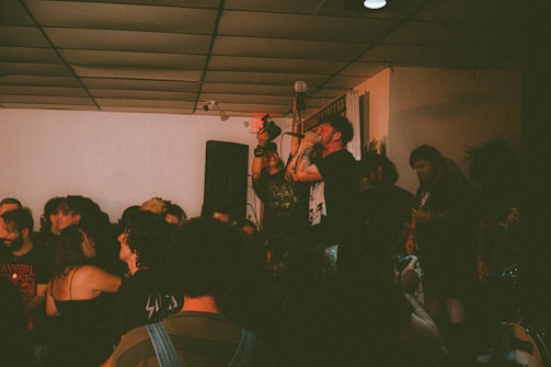 A live music performance is taking place in a compact indoor venue with a crowd of people gathered around. The lead singer is passionately holding a microphone and singing, surrounded by audience members who appear engaged and energetic. The setting is dimly lit, creating an intimate ambiance.